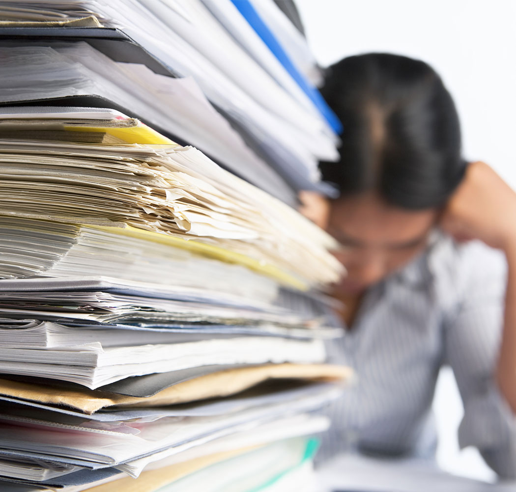 woman holding her head in her hands looking exhausted sitting with giant pile of paperwork