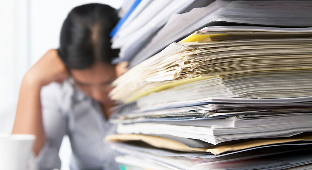 woman-with-stack-of-paper overwhelmed looking businesswoman with a giant stack of papers on her desk